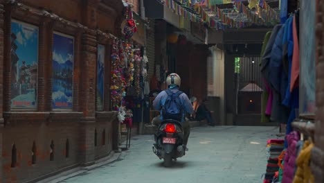 A-person-riding-a-motorbike-passing-through-a-narrow-street-with-prayer-flags-hanging-from-buildings,-in-Kathmandu,-Nepal