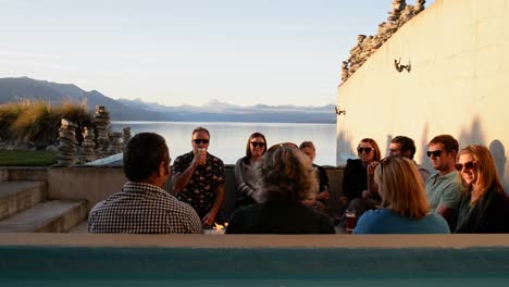 Group-of-happy-people-sitting-around-fire-on-picturesque-lakeview-patio-near-Mt-Cook