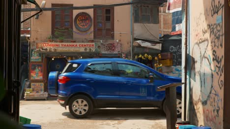 Ein-Blaues-Auto,-Das-Durch-Eine-Schmale-Straße-Mit-Kleinen-Geschäften-Und-Märkten-In-Kathmandu,-Nepal,-Fährt