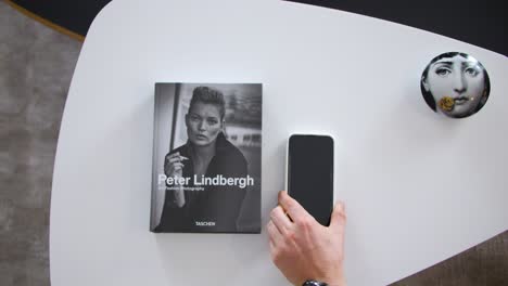 view-from-above-of-a-small-white-designer-coffee-table-on-which-a-book-with-a-woman-in-black-and-white-on-the-cover,-a-human-hand-places-a-smartphone-beside-it,-blurred-gray-floor