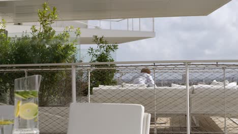 Cóctel-De-Verano-En-Primer-Plano-Sobre-Una-Moderna-Mesa-Blanca-Al-Aire-Libre,-Luego-Un-Hombre-Detrás-Caminando-Hacia-La-Derecha-Y-Sentado-En-Su-Moderno-Sofá-Blanco-Disfrutando-De-La-Vista-De-La-Ciudad-Desde-Su-Terraza