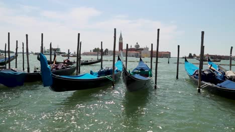 Gondolas-docking-in-a-pier-with-gondolier-and-boats-passing-by-and-the-bell-tower-of-St