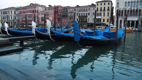 Venetian-gondolas-docking-in-a-Grand-Canal's-pier-with-tourist-boat-passing-in-the-background-in-Venice,-Italy