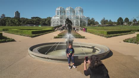 Touristen-Besuchen-Den-Botanischen-Garten-Von-Curitiba,-Im-Vordergrund-Die-Statue-Der-„mütterlichen-Liebe“-Und-Im-Hintergrund-Der-„Kristallpalast“,-Zeitrafferaufnahme