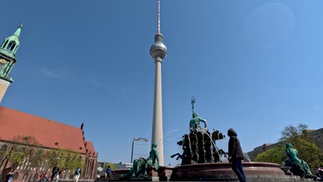 Low-Angle-Shot-Of-The-Neptune-Fountain-In-Berlin-and-Berliner-Fernsehturm-Against-Clear-Blue-Skies-With-People-Walking-Past