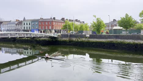 Ruderer-Paddelt-Durch-Das-Grüne-Wasser-Des-Flusses-Lee-In-Cork-City-Mit-Blick-Auf-Stadthäuser-Und-Eine-Brücke