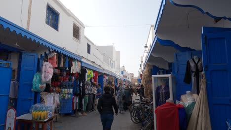People-walking-along-Picturesque-bazaar-street-with-blue-painted-doors-in-Essaouira-Medina,-Morocco