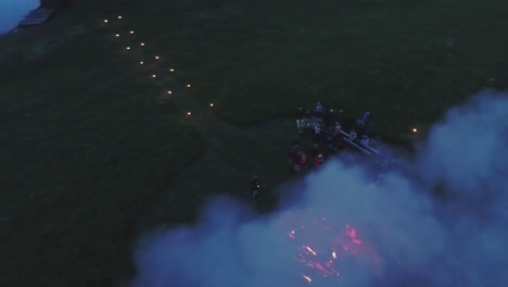 People-Enjoy-Massive-Bonfire-At-Night-in-a-meadow-field,-Aerial-Fly-Away-View