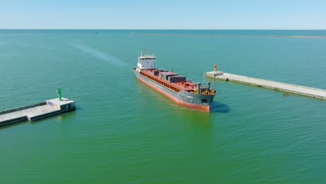 Aerial-establishing-view-of-large-red-cargo-ship-entering-Port-of-Liepaja-,-Karosta-bridge,-sunny-day,-calm-Baltic-sea,-city-in-background,-wide-drone-shot-moving-backward