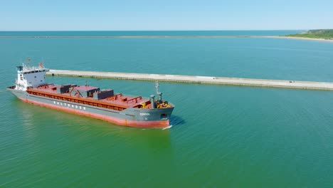 Aerial-establishing-view-of-large-red-cargo-ship-entering-Port-of-Liepaja-,-Karosta-bridge,-sunny-day,-calm-Baltic-sea,-city-in-background,-wide-drone-shot