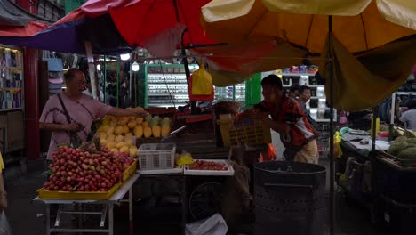Vista-De-Vendedores-De-Frutas-Que-Venden-Frutas-Frescas-Y-Un-Cliente-Que-Ofrece-Una-Barra-De-Cigarrillo-En-Petaling-Street,-Kuala-Lumpur,-Malasia