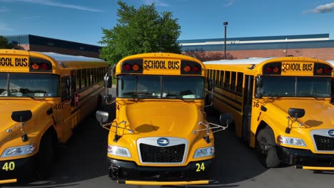 Toma-Aérea-Ascendente-De-Autobuses-Escolares-Amarillos-Estacionados-Frente-Al-Edificio-De-Una-Escuela-Pública-Estadounidense