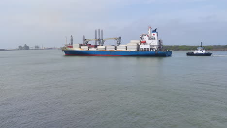 A-massive-container-ship-commands-the-bay-as-it-gracefully-approaches-an-industrial-port,-with-a-dedicated-tug-boat-following-closely-behind-scenic-view,-Industrial-port,-logistic,-Container-ship