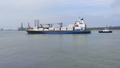 A-massive-container-ship-commands-the-bay-as-it-gracefully-approaches-an-industrial-port,-with-a-dedicated-tug-boat-following-closely-behind