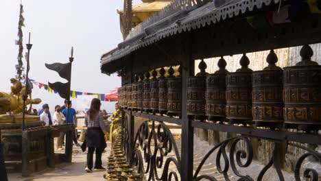 Row-Of-Prayer-Wheels-At-Temple-In-Kathmandu-With-Tourist-Walking-Past-And-Monkey-Jumping-Out
