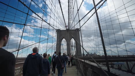 Menschen,-Die-über-Die-Brooklyn-Bridge-Laufen,-Mit-Blauem-Himmel-Und-Wolken,-Die-Durch-Aufhängedrähte-Zu-Sehen-Sind