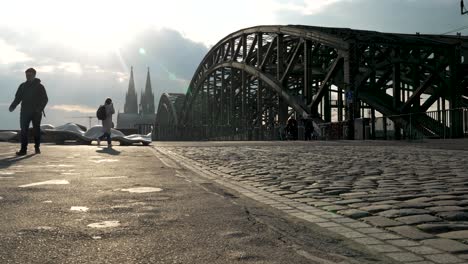 Wunderschönes-Kopfsteinpflaster-Mit-Der-Hohenzollernbrücke-Und-Dem-Kölner-Dom,-Der-In-Sonnenlicht-Getaucht-Ist,-Wenn-Menschen-Vorbeigehen