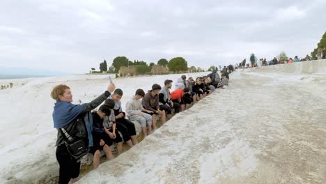 Group-of-people-sit,-bathe-feet-and-enjoy-natural-hot-spring-visitor-attraction-at-Pamukkale-Turkey