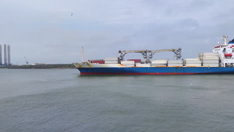 Ein-Riesiges-Containerschiff-Beherrscht-Die-Bucht,-Während-Es-Sich-Elegant-Einem-Industriehafen-Nähert,-Dicht-Gefolgt-Von-Einem-Speziellen-Schlepper,-Der-Sich-Langsam-In-Richtung-Hafen-Bewegt,-Um-Container-Und-Logistik-Zu-Entladen