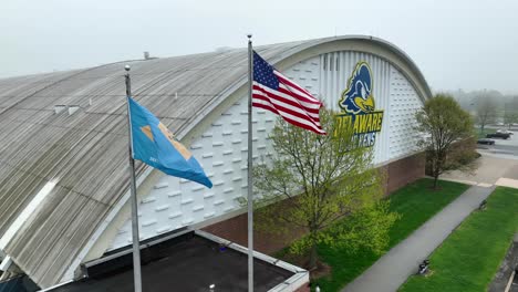 Aerial-shot-of-American-and-Delaware-flags-waving-in-front-of-University-of-Delaware-Blue-Hens-field-house