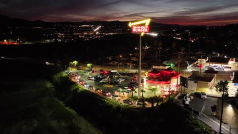 Toma-Aérea-De-La-Cadena-De-Comida-Rápida-De-La-Costa-Oeste,-In-n-Out-Burger,-Manejando-Una-Larga-Fila-De-Clientes-De-Autoservicio-Mientras-El-Día-Se-Convierte-En-Noche-En-Una-Ubicación-Del-Sur-De-California-En-Santa-Clarita,-California,-Junto-A-La-Autopista-14.