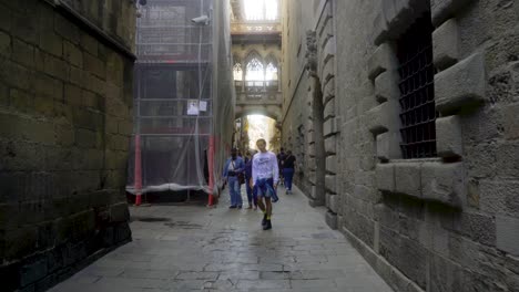 Pont-del-Bisbe,-Barcelona,-with-couple-and-tourists-walking-through-narrow-street-in-Barri-Gothic-Quarter