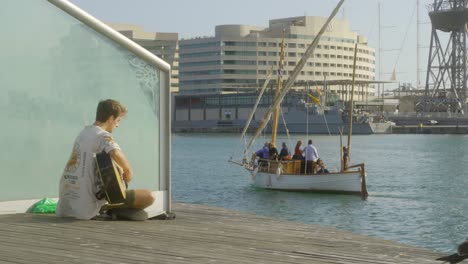 Young-blond-man-playing-the-guitar-during-sunset-by-the-water-at-the-commercial-harbour-of-Barcelona,-Spain