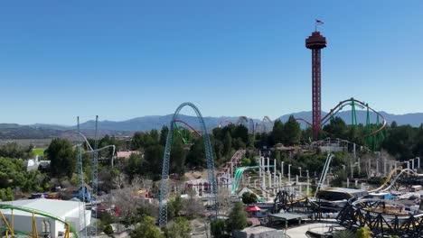 Aerial-slow-motion-of-guests-riding-rollercoasters-at-the-Six-Flags-Theme-Park-in-Santa-Clarita,-California-as-traffic-passes-on-the-5-freeway-and-hills-are-seen-in-the-background