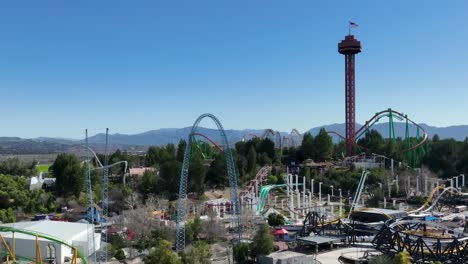 Cámara-Lenta-Aérea-De-Invitados-Que-Viajan-En-Montañas-Rusas-En-El-Parque-Temático-Six-Flags-En-Santa-Clarita,-California,-Mientras-El-Tráfico-Pasa-Por-La-Autopista-5-Y-Se-Ven-Colinas-Al-Fondo.