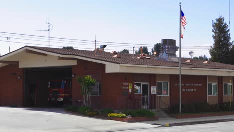 Toma-Estática-De-Una-Estación-De-Bomberos-Del-Condado-De-Los-Ángeles-Con-Banderas-Ondeando-En-La-Brisa-Y-El-Tráfico-Que-Pasa.