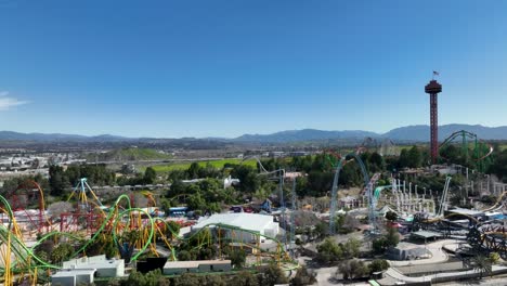Aerial-ascent-showing-guests-riding-many-rollercoasters-and-Six-Flags-Theme-Park-in-Santa-Clarita,-California-with-Interstate-5-freeway-in-and-surrounding-hills-in-the-background-on-a-clear-sunny-day