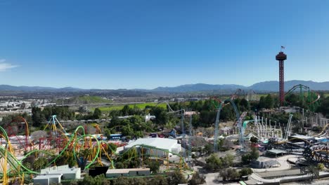 Ascenso-Aéreo-Que-Muestra-A-Los-Visitantes-Montando-Muchas-Montañas-Rusas-Y-El-Parque-Temático-Six-Flags-En-Santa-Clarita,-California,-Con-La-Autopista-Interestatal-5-Y-Las-Colinas-Circundantes-Al-Fondo-En-Un-Día-Claro-Y-Soleado.