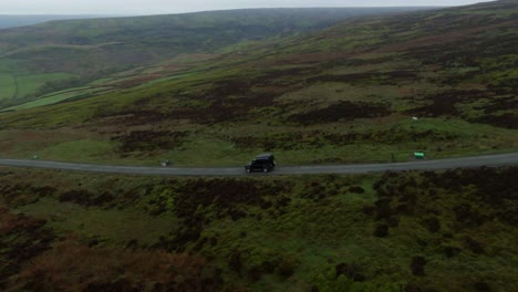 Drone-rise-of-Black-Historic-Land-Rover-Defender-driving-down-winding-thin-single-lane-alongside-huge-green-valleys-and-hills-in-the-North-Yorkshire-Moors-near-Rosedale