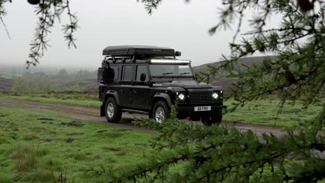 Schwarzer-Modifizierter-Camping-Landrover-Verteidiger-Mit-Dachzelt,-Der-An-Einem-Bewölkten,-Launischen-Tag-Statisch-Auf-Einer-Einspurigen-Landstraße-Geparkt-Ist-Und-Bäume-Und-Grün-Als-Vordergrund-Verwendet