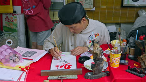 A-calligraphy-master-creates-beautiful-handwriting-using-penmanship-techniques-on-a-red-table,-surrounded-by-his-tools,-against-a-penmanship-background