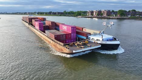 Aerial-View-Off-Forward-Bow-Of-Arctic-Cargo-Container-Ship-Paired-With-Barge-Travelling-Along-River-Noord