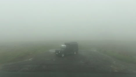 Drone-tilt-up-of-black-Land-Rover-Defender-parked-in-very-cloudy-and-misty-mountain-hills-with-zero-visibility-and-fog-lights-shining