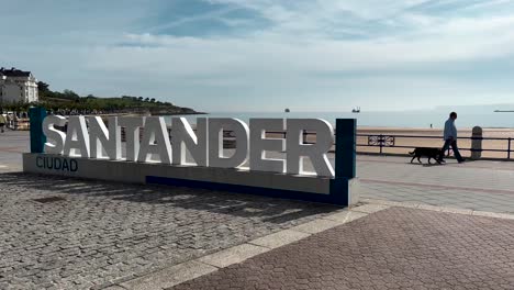 Nahaufnahme-Des-Santander-Schildes-Vor-Der-Promenade-Mit-Sandstrand-Und-Meer-An-Sonnigen-Tagen,-Zeitlupe