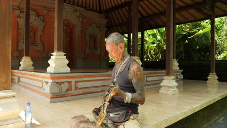 Toma-De-Paralaje-En-Cámara-Lenta-De-Un-Encantador-De-Serpientes-Tradicional-En-Un-Templo-Balinés-Que-Lleva-Una-Serpiente-Alrededor-Del-Cuello-Y-La-Controla-Elegantemente-En-Bali,-Indonesia.