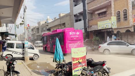 Autobús-Rosa-Pasando-Por-Un-Mecánico-De-Automóviles-Trabajando-En-La-Puerta-Al-Lado-De-La-Carretera-En-Saddar,-Karachi-Con-Aire-Polvoriento-En-El-Fondo