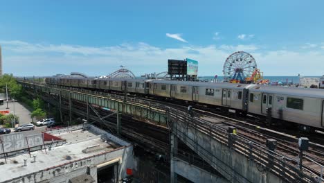 Aerial-shot-of-NYC-commuter-train-running-on-tracks-in-front-of-Coney-Island-amusement-park