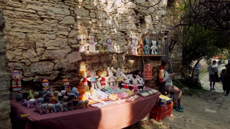 Frau-Verkauft-Handgefertigte-Türkische-Puppen-Auf-Einer-Gepflasterten-Straße-Im-Schatten-Einer-Steinmauer