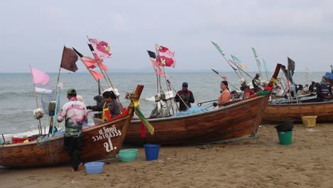 Hölzerne-Fischerboote,-Windpeitschende-Flaggen-Und-Fischer,-Die-Netze-Vorbereiten,-Während-Sie-Frische-Meeresfrüchte-Am-Strand-In-Jomtien,-Thailand,-Verkaufen