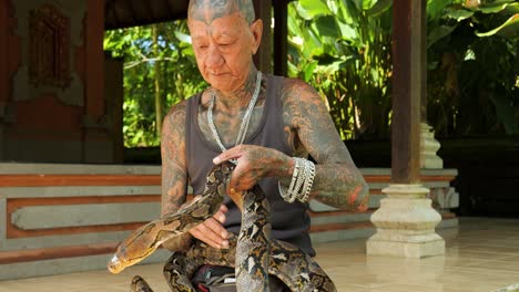 Toma-De-Paralaje-En-Cámara-Lenta-De-Un-Domador-De-Serpientes-Tradicional-En-Un-Templo-Balinés-Que-Lleva-Una-Serpiente-Alrededor-Del-Cuello-Y-La-Controla-Elegantemente-En-Bali,-Indonesia.
