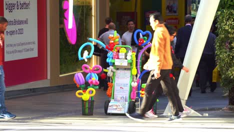 Close-up-shot-of-a-busking-balloon-twister-stall-at-bustling-downtown-Brisbane-city,-Queen-street-mall-with-large-crowds-of-passerby-on-the-street-on-a-sunny-day