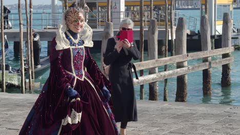 Venecia,-Italia---13-De-Febrero-De-2023-Mujer-Con-Vestido-Medieval-Antiguo-Y-Máscara-Caminando-Por-Calles-Concurridas-Siendo-Fotografiada-Por-Un-Turista