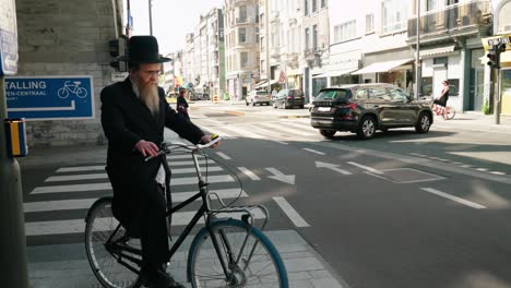 Orthodox-Jew-on-bike-waiting-at-the-intersection-to-cross-the-city-street-in-the-Antwerp's-diamond-district,-Belgium---Medium-angle