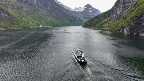 Tourist-boat-full-of-passengers-and-cars-are-cruising-the-Geiranger-Fjord-in-Norway---Forward-moving-aerial-overtaking-the-ship-with-the-eagles-road-to-the-left-and-Geiranger-town-in-background