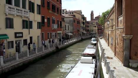 Touristen,-Die-Auf-Einem-Schmalen-Pfad-Neben-Traditionellen-Venezianischen-Häusern-Neben-Einem-Schmalen-Kanal-In-Venedig-Spazieren