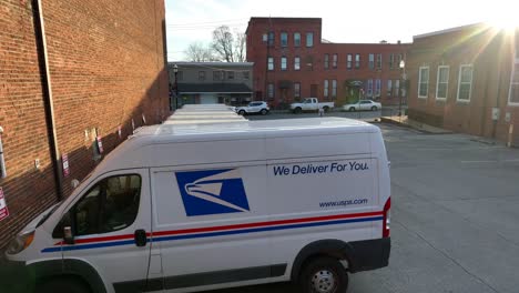 Drone-view-of-a-mail-delivery-van-in-the-parking-lot-of-a-post-office-with-the-setting-sun-gleaming-over-adjacent-buildings